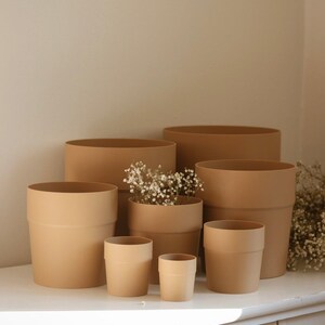 May include: A collection of seven cylindrical plant pots in a solid matte tan colour, arranged in varying heights and widths. One of the central pots contains a small cluster of dried white baby's breath flowers, adding a delicate texture.