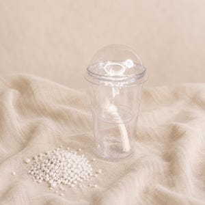 May include: A clear plastic cup with a dome lid and a white wick inside, accompanied by a small pile of white granular material. The cup features a transparent design with a visible internal structure and a central white wick extending upwards.