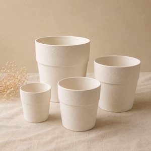 May include: A set of four off-white plant pots in varying sizes, each featuring a textured, matte surface and a simple cylindrical shape with a slightly wider rim at the top. The pots are arranged in a cluster, showcasing their uniform colour.