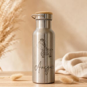 Könnte beinhalten: Eine Edelstahl-Wasserflasche mit Holzdeckel und silberfarbenem Griff. Die Flasche zeigt eine schwarze Illustration eines Zebras und den Namen "Angsar" in Schreibschrift. Für wiederverwendbare Flüssigkeitszufuhr.
