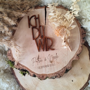 Op de afbeelding: Een houten trouwteken met de woorden "ICH DU WIR" in bruine letters. De namen "Patric & Nele" en de datum "18.SEPTEMBER 2025" zijn eronder gegraveerd. Het teken is versierd met gedroogde bloemen en ligt op een houten schijf.