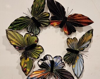 May include: A collection of six decorative paper butterflies arranged in a circle. The butterflies have black bodies and wings in various colors, including green, orange, and blue. Each butterfly has a textured, pleated wing design.