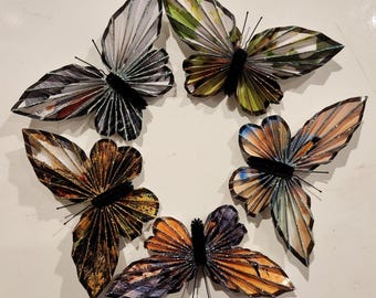 May include: Five paper butterflies with glitter accents in various colors, including green, brown, blue, and orange, arranged in a circle on a white background.