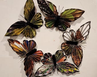 May include: Six paper butterflies with different patterns and colors, including green, brown, black, and white. The butterflies are arranged in a circle on a white background.