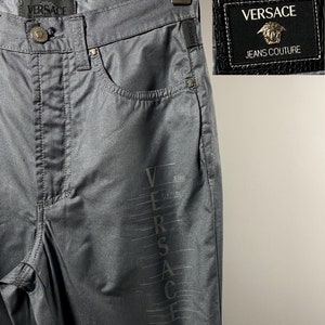 Puede incluir: Vaqueros de denim gris oscuro con un logotipo vertical plateado 'VERSACE JEANS COUTURE' en la pierna derecha. Los vaqueros tienen un estilo clásico de cinco bolsillos y cierre de botón.
