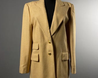 May include: A tan and white houndstooth blazer with two front pockets and gold buttons. The blazer is displayed on a black mannequin and has a classic, tailored design. The jacket is a neutral color and suitable for various occasions.
