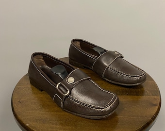 Pode incluir: Um par de mocassins de couro marrom com um detalhe de fivela. Os mocassins estão em cima de um banquinho de madeira.