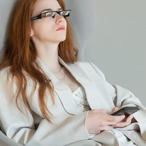 May include: Black prism glasses with a rectangular frame are worn by a person. The person is wearing a white blazer and holding a smartphone. The glasses are designed to allow the wearer to read while lying down.