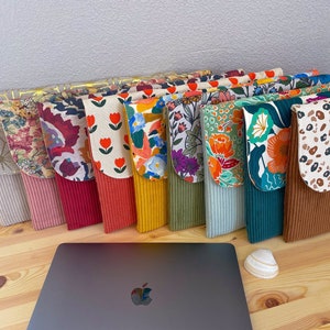 Peut inclure: Une collection de 12 housses pour ordinateur portable colorées avec différents motifs. Les housses sont en velours côtelé et ont une fermeture à rabat. Les housses sont doublées d'un tissu doux. Les housses sont disposées en rangée sur une surface en bois.