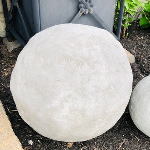 May include: Two light gray, spherical garden stones of different sizes rest on dark mulch. A large, square, dark gray metal planter with a floral accent is in the background, along with green foliage.
