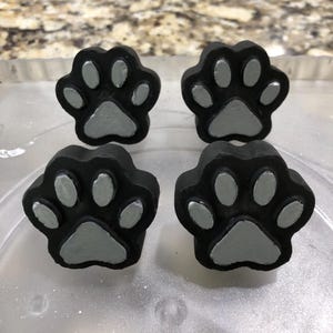 May include: Four black paw print shaped cabinet knobs with silver-colored pads. The knobs are arranged in a square pattern, perfect for a pet-themed room or furniture update. Each knob has a detailed paw print design.