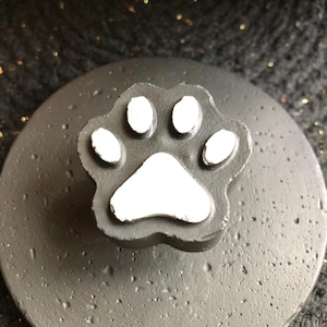 May include: A dark gray, round object with a raised paw print design. The paw print is black with white pads. The object has a textured surface and is likely a decorative item or a lid.