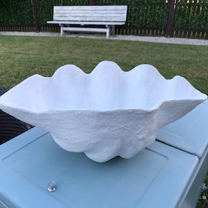 May include: A large, white, clam-shaped decorative bowl with a textured surface. The bowl is sitting on a light blue surface, with a grassy lawn and a white bench in the background. The bowl's wavy edges give it a natural, organic look.