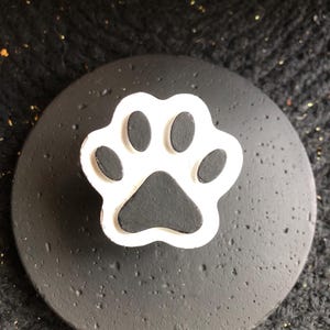 May include: A black, round lid with a white and black paw print design. The paw print is raised and has a white outline. The lid has a textured surface.