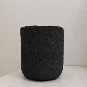 May include: A cylindrical vessel with a deep black color and a highly textured, rough, matte surface. The object features a simple, straight-sided design with a rounded base and an open top, presenting a uniform appearance throughout its exterior.