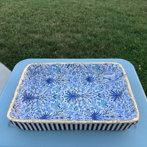 May include: Rectangular decorative tray with a floral pattern in shades of blue and white. The tray has a gold trim and black and white striped sides. The tray is sitting on a light blue surface.