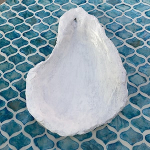 May include: A white, oyster shell-shaped decorative dish. The dish is painted white and sits on a blue and white patterned surface. The shell has a rough, textured appearance.