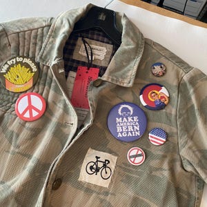 May include: Camouflage-patterned jacket with various pin-on buttons. Buttons include a peace sign, a fries graphic, a bicycle, and political campaign buttons. The jacket has a plaid lining and a tag.