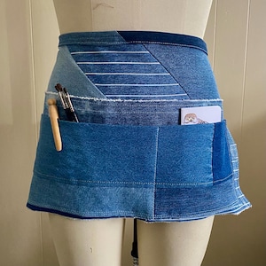 May include: A denim apron with multiple pockets, perfect for artists. The apron is constructed from various shades of blue denim, with a patchwork design. It features pockets holding paintbrushes, a pen, and a small card with an owl illustration. The apron is displayed on a mannequin.