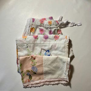 May include: A stack of vintage fabric pieces, including a pair of white floral print shorts with a drawstring waist. Other pieces feature floral and botanical patterns in pink, blue, and green, with a lace trim.