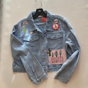 May include: Light blue denim jacket with multiple decorative patches. Patches include a cactus, peace sign, and the words "Mojave Desert". The jacket has a button-up front, two chest pockets, and long sleeves.