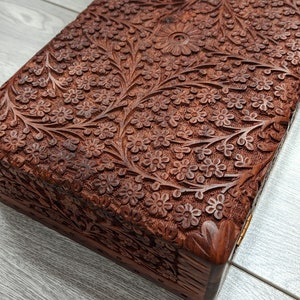 Puede incluir: Una caja de madera con intrincados tallados florales. La caja est&aacute; hecha de madera marr&oacute;n oscura y tiene una tapa con bisagras. La caja est&aacute; cerrada y se encuentra sobre una superficie de madera gris claro.