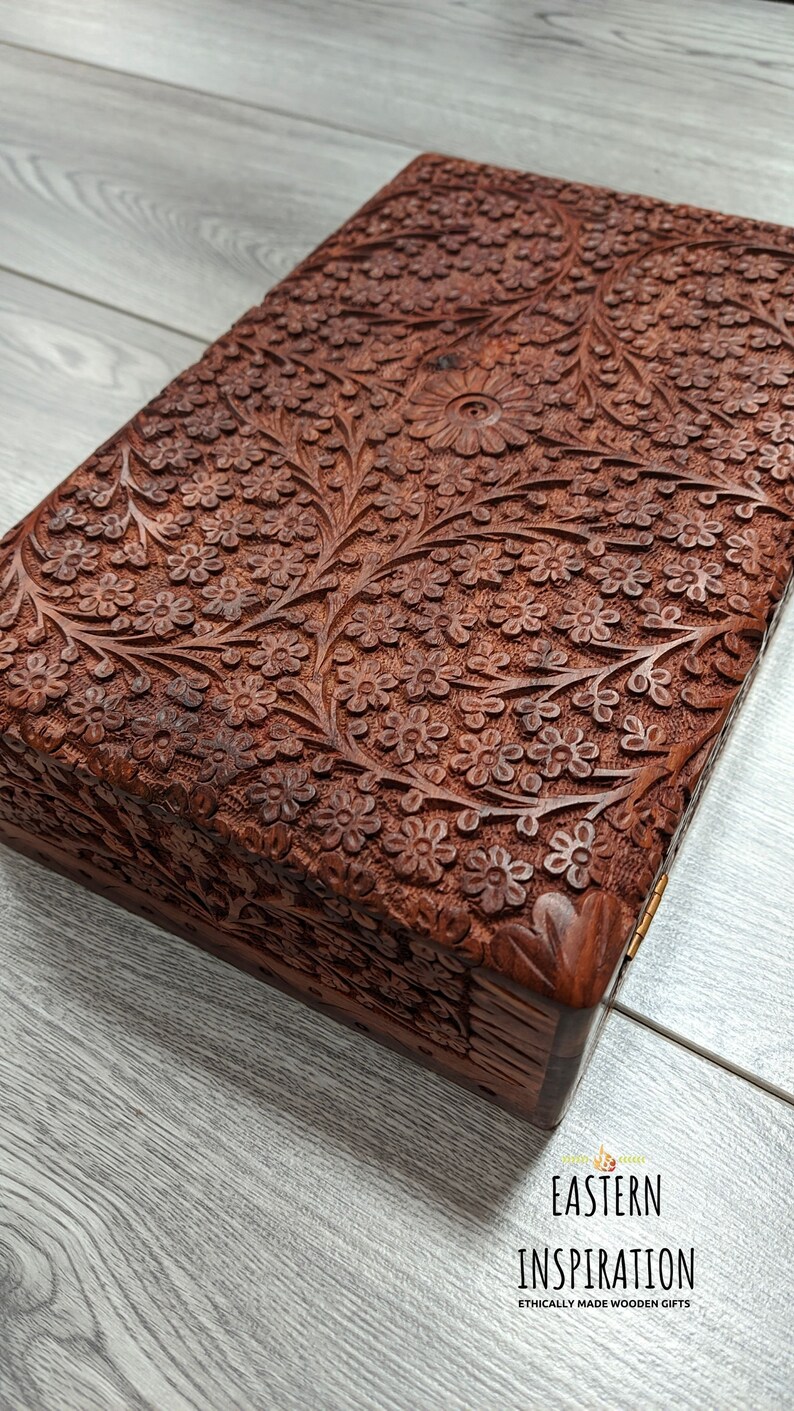 Puede incluir: Una caja de madera con intrincados tallados florales. La caja est&aacute; hecha de madera marr&oacute;n oscura y tiene una tapa con bisagras. La caja est&aacute; cerrada y se encuentra sobre una superficie de madera gris claro.