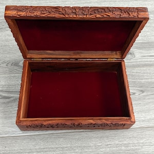 Puede incluir: Una caja de madera con intrincados tallados en la tapa y los lados. La caja est&aacute; abierta y forrada con terciopelo rojo. La caja est&aacute; hecha por Eastern Inspiration, una empresa que fabrica regalos de madera hechos &eacute;ticamente.
