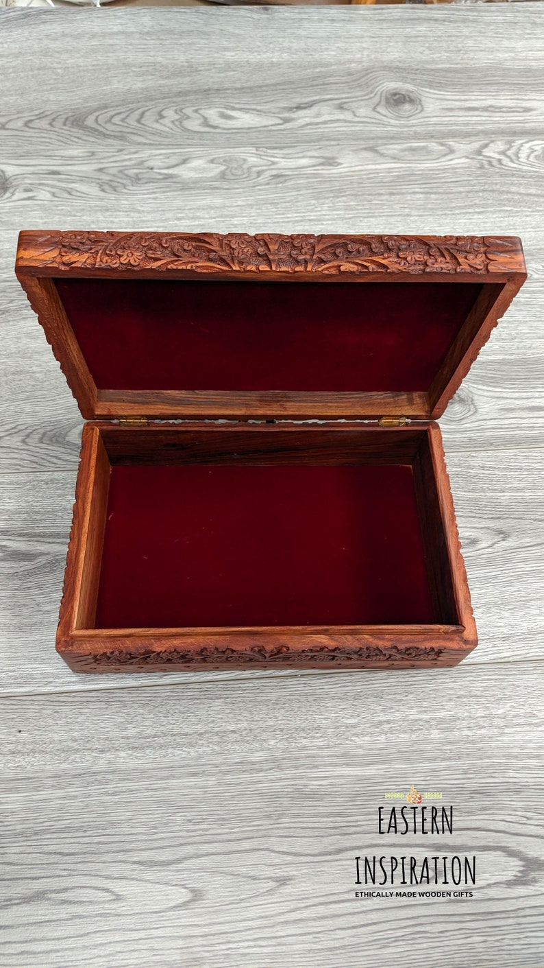 Puede incluir: Una caja de madera con intrincados tallados en la tapa y los lados. La caja est&aacute; abierta y forrada con terciopelo rojo. La caja est&aacute; hecha por Eastern Inspiration, una empresa que fabrica regalos de madera hechos &eacute;ticamente.