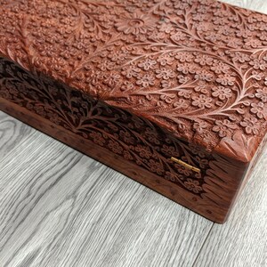 Puede incluir: Una caja de madera con intrincados tallados florales. La caja est&aacute; cerrada y tiene una bisagra de lat&oacute;n. La caja est&aacute; hecha de madera oscura y tiene un tono marr&oacute;n rojizo.