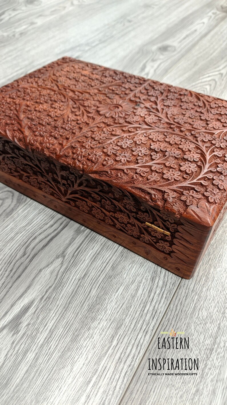 Puede incluir: Una caja de madera con intrincados tallados florales. La caja est&aacute; cerrada y tiene una bisagra de lat&oacute;n. La caja est&aacute; hecha de madera oscura y tiene un tono marr&oacute;n rojizo.