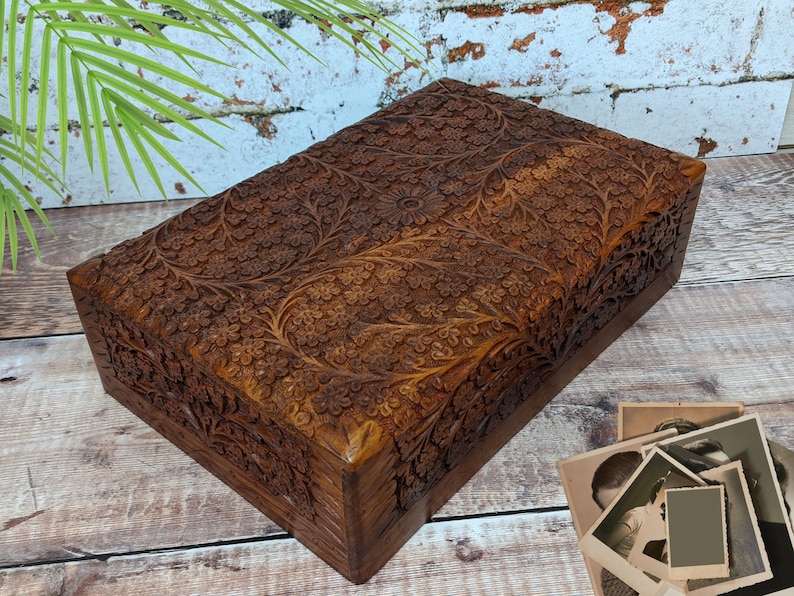 Puede incluir: Una caja de madera con intrincados tallados florales. La caja est&aacute; hecha de madera marr&oacute;n oscura y tiene una forma rectangular. Los tallados son detallados y cubren toda la superficie de la caja.