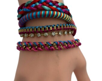 Puede incluir: Un conjunto de pulseras trenzadas de colores con una cadena de oro y una pulsera de diamantes de imitación. Las pulseras están apiladas en la muñeca de una persona.
