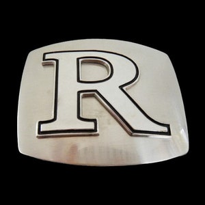 May include: A silver-colored belt buckle with a large, capital letter "R" in the center. The letter is outlined in black, and the buckle has a brushed metal finish. The buckle is rectangular with rounded corners.