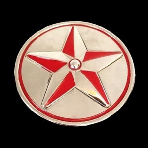 May include: A circular, silver-coloured emblem featuring a red star at its centre. The star has five points and is encircled by a red border. A small, silver-coloured circle is in the star's centre.