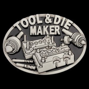 Op de afbeelding: Een zilveren gesp met een zwarte achtergrond. De gesp heeft de tekst "Tool & Die Maker" en een gedetailleerd beeld van een gereedschap- en matrijsset.
