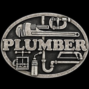 Op de afbeelding: Ovaalvormige, zilverkleurige gesp met het woord "PLUMBER" in vette letters. De gesp toont diverse loodgietersgereedschappen, waaronder een moersleutel, pijpsnijder en een brandstoftank, allemaal in een gedetailleerd, reliëf ontwerp.