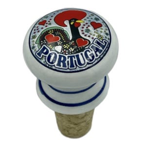 Puede incluir: Un tapón de botella de cerámica blanca con base de corcho. La parte superior presenta una colorida ilustración de un gallo con la palabra "PORTUGAL" en azul. El gallo tiene una cresta roja y detalles en forma de corazón.