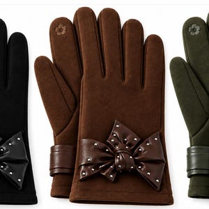 Könnte beinhalten: Drei Paar Handschuhe in Schwarz, Braun und Olivgrün. Jedes Paar hat eine glatte Oberfläche, eine dekorative Schleife am Handgelenk mit kleinen silbernen Nieten und ein kleines blumenförmiges Detail am Zeigefinger für Touchscreen-Bedienung.