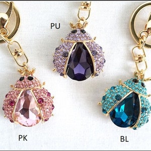 May include: Three ladybug keyrings in pink, purple, and blue. Each ladybug features a large, faceted gem body and is encrusted with smaller crystals. The keyrings have gold-coloured metal hardware. The letters "PK", "PU", and "BL" are visible.
