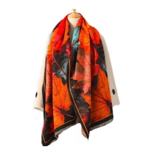 Puede incluir: Una bufanda vibrante con un gran estampado de hojas en tonos naranja, rojo y verde azulado sobre un fondo negro. La bufanda está drapeada sobre un maniquí, mostrando su diseño y textura. La bufanda tiene un borde negro.