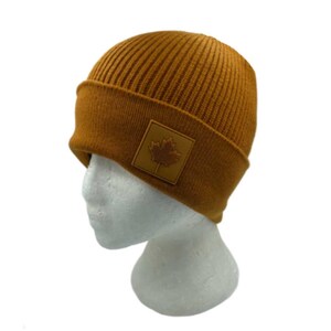 Op de afbeelding: Een bruine gebreide beanie met een bruine leren patch met een esdoornblad.