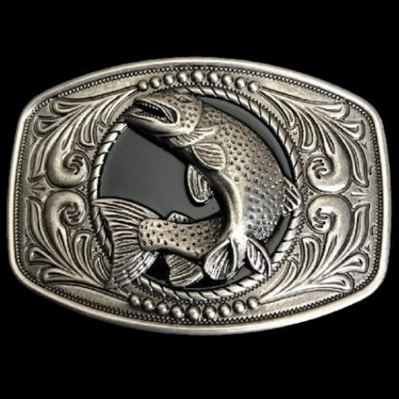 May include: Silver belt buckle with an ornate design featuring a jumping fish in the center. The fish is detailed with scales and fins, and the buckle has a black background.