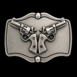 May include: A silver-colored metal belt buckle featuring two revolvers facing each other. The revolvers have detailed grips with star accents. The buckle has a decorative border and a dark background.