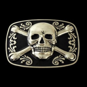 May include: A rectangular metal belt buckle featuring a skull with crossed bones. The skull is centered, with detailed teeth and eye sockets. The background is black, and the buckle has ornate scrollwork.