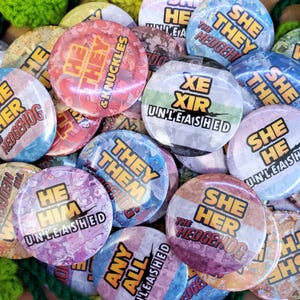 May include: A collection of circular pin-back buttons featuring various pronoun combinations and text such as He They & Knuckles, Xe Xir Unleashed, They Them, She He Unleashed, He Him Unleashed, She Her The Hedgehog, and Any All Unleashed.