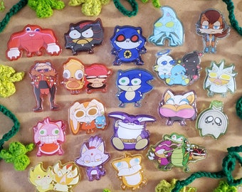 Puede incluir: Una colección de pines acrílicos con varios personajes de la franquicia Sonic the Hedgehog. Los pines muestran personajes como Sonic, Shadow y otros en colores vibrantes y diseños detallados. Los pines están dispuestos sobre una superficie de madera con vides y hojas de ganchillo verdes.