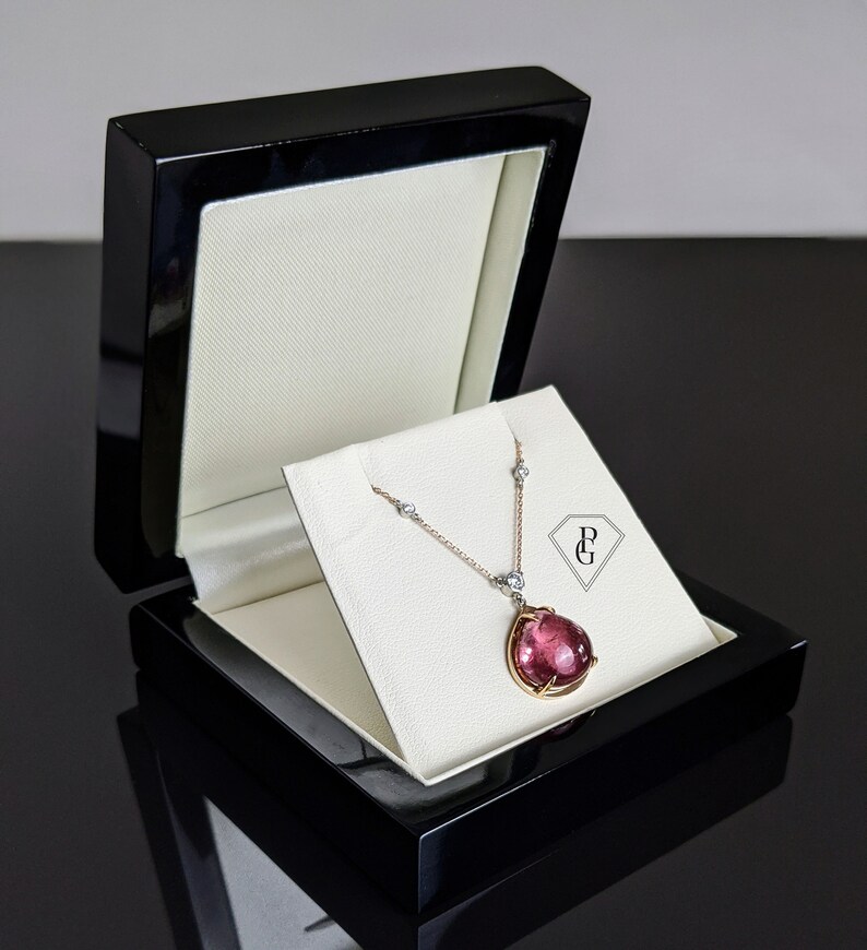 Op de afbeelding: Een ketting met een roze edelsteen hanger, een delicate ketting en kleine diamant accenten. De ketting wordt gepresenteerd in een zwarte fluwelen doos met de initialen "P & G" aan de binnenkant.