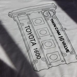 Könnte beinhalten: Weißes T-Shirt mit einer schwarzen Linienzeichnung eines Toyota 1600 Motors. Die Zeichnung enthält den Text "Twin Cam 16 Valve" und "Engine Oil".
