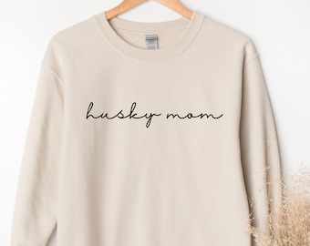 Op de afbeelding: Een beige sweatshirt met de woorden "husky mom" in zwart script. Het sweatshirt hangt aan een houten hanger. Gedroogd siergras staat rechts van de sweatshirt.